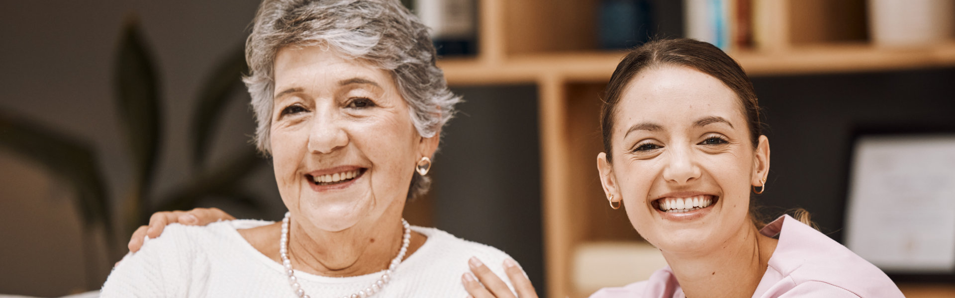 A young woman and a elder woman smile together in a friendly moment, inside a home